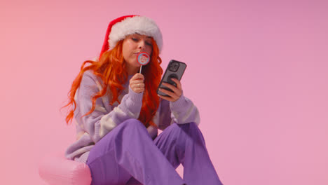 Studioaufnahme-Einer-Jungen-Frau-Der-Generation-Z-Mit-Weihnachtsmütze,-Die-Süßigkeitenlutscher-Isst-Und-Auf-Ihr-Mobiltelefon-Schaut-2
